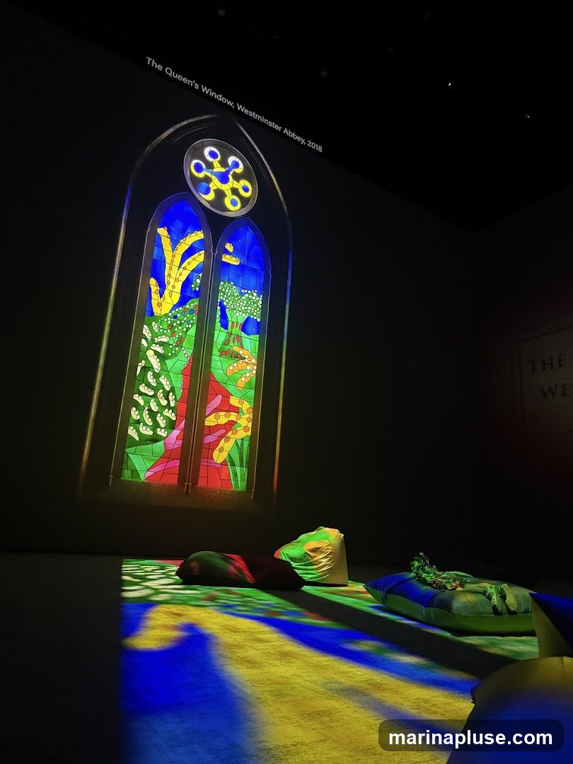 A projection of a stained-glass window titled "The Queen's Window, Westminster Abbey, 2018" is displayed on a dark wall. The window features vibrant colors and abstract designs. In the foreground, colorful cushions are scattered on the floor, illuminated by the projected light.