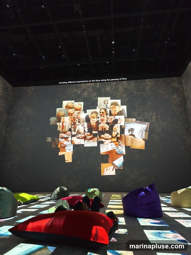 An art installation featuring a projection of photographs of faces, arranged in a collage on a dark wall. Text above the projection reads "showing different expressions on the face using the passing of time." The floor is illuminated with colorful projected squares, and several beanbag chairs are scattered around the room, with a person lying on one of them.