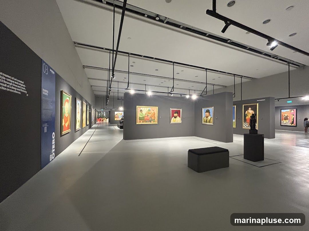An art gallery exhibition hall with paintings displayed on the walls. The hall is spacious with a grey floor and dark grey walls. Track lighting illuminates the artwork. Several benches are placed throughout the hall for visitors. In the foreground, a large dark grey bench is visible. Further down the hall, more paintings and a sculpture are displayed. A sign on the left wall reads '03 BOTERO PAINTED DRAMA'.