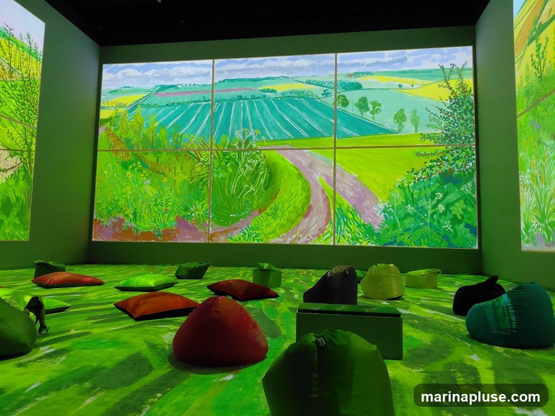 An immersive art exhibition featuring large-scale projections of a vibrant landscape painting. The room is filled with various beanbag chairs and cushions scattered on the floor, inviting visitors to relax and enjoy the visual experience. The projections cover the walls, creating a 360-degree environment.