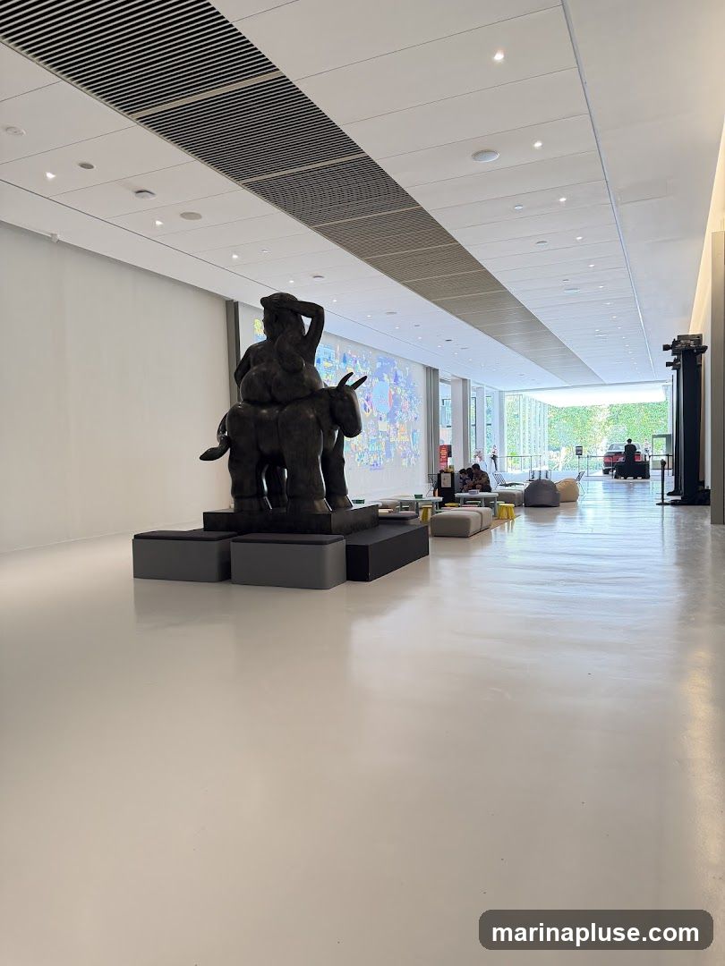 A wide, brightly lit hallway with a polished white floor. In the foreground, a large, dark bronze sculpture depicts figures riding a donkey. The sculpture is placed on a black and grey platform with seating. In the background, a colorful mural is projected onto a wall, and several people are seated on beanbag chairs and sofas in a lounge area. The hallway leads to an open, sunlit space with greenery visible outside.