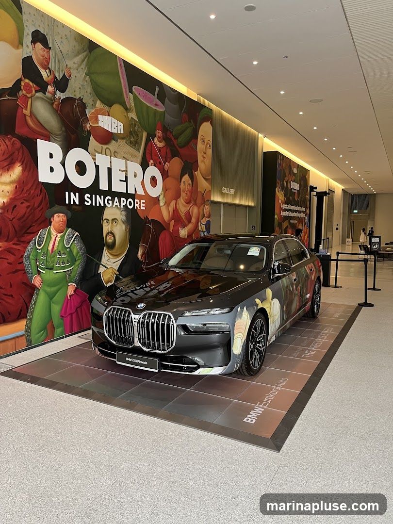 A black BMW 7 Series car is displayed on a tiled platform in front of a large mural featuring artwork by Botero. The mural has the text "BOTERO IN SINGAPORE" prominently displayed. The car has custom artwork on its side, inspired by the Botero style. The background shows a modern interior with a gallery sign and lighting.