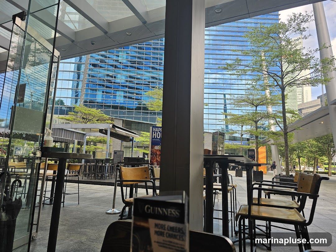 An outdoor seating area with tables and chairs is visible, with a large modern building with a blue glass facade in the background. Trees and greenery are interspersed throughout the scene. A sign for Guinness is partially visible in the foreground.