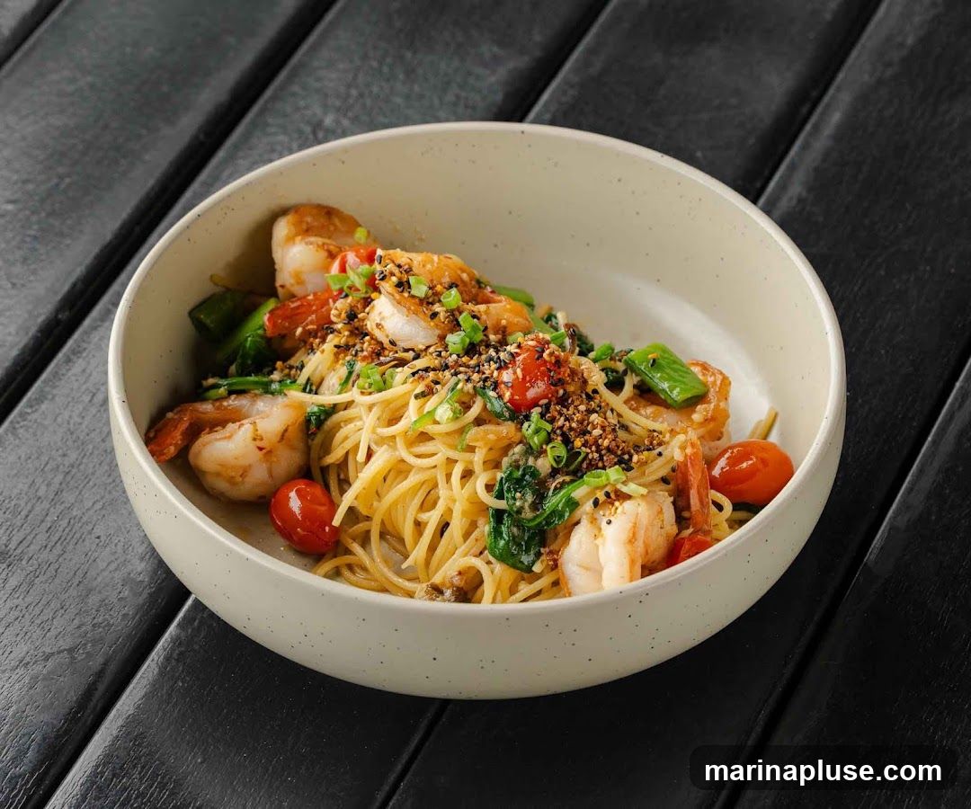 A close-up shot of a bowl of spaghetti with shrimp, cherry tomatoes, and greens. The dish is garnished with chopped scallions and a sprinkle of seeds. The bowl is placed on a dark wooden surface.