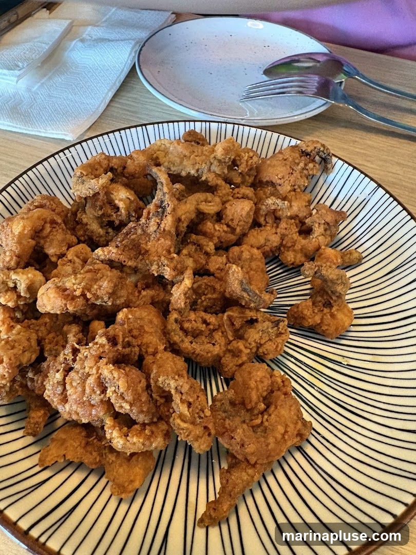 A plate of golden-brown fried calamari is served on a plate with a black and white striped pattern. A white plate with cutlery and napkins are visible in the background.