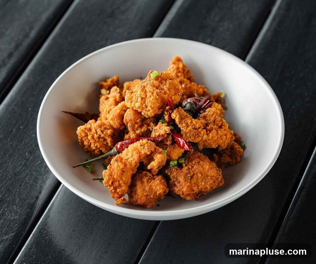 A white bowl filled with golden-brown, crispy fried chicken bites, garnished with chopped green onions and dried red chilies, served on a dark wooden surface.