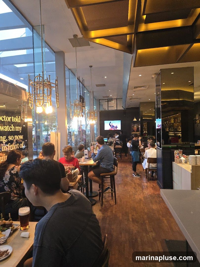 The image shows the interior of a restaurant with several patrons seated at tables. The decor features large windows, modern lighting fixtures resembling trumpets, and wooden flooring. A television screen is visible in the background, displaying an image. Staff members are also seen attending to customers.