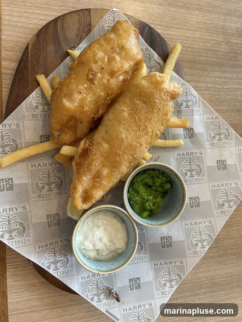 A serving of fish and chips on a wooden board, lined with paper featuring the logo 'HARRY'S EST. 1992'. The dish includes two pieces of battered fish, a portion of french fries, mushy peas in a small bowl, and a creamy sauce in another small bowl. A lemon wedge is also visible.