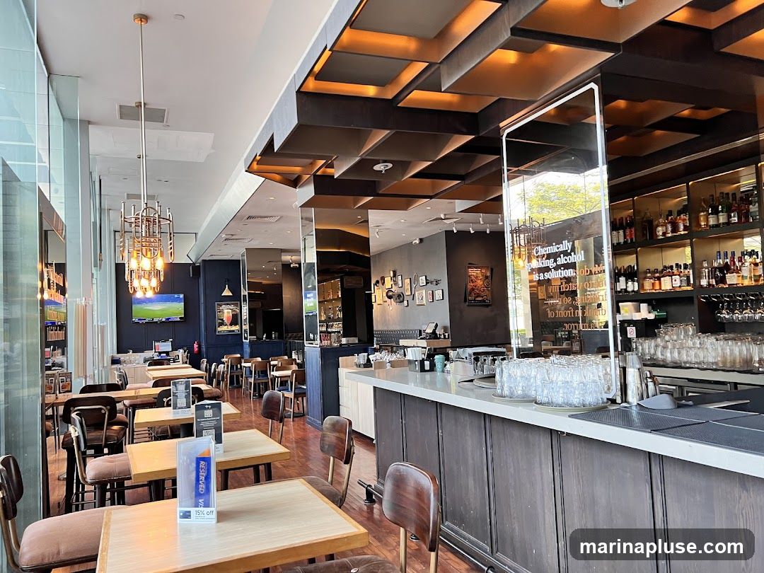 The interior of a bar with tables and chairs, a bar counter, and shelves stocked with bottles. A television screen is visible on the wall. A glass partition on the bar has text that reads "Chemically speaking, alcohol is a solution."
