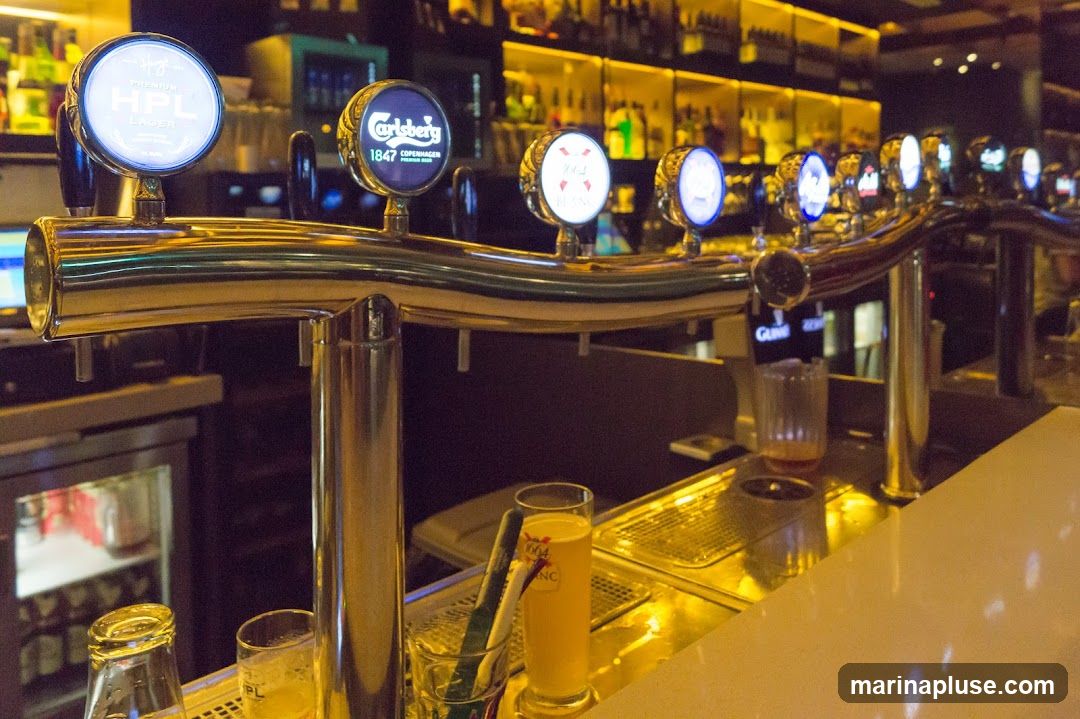 A row of illuminated beer taps with various logos, including HPL, Carlsberg, and Guinness, are visible at a bar. In the foreground, there are glasses and a bottle. The background shows shelves stocked with bottles of alcohol.