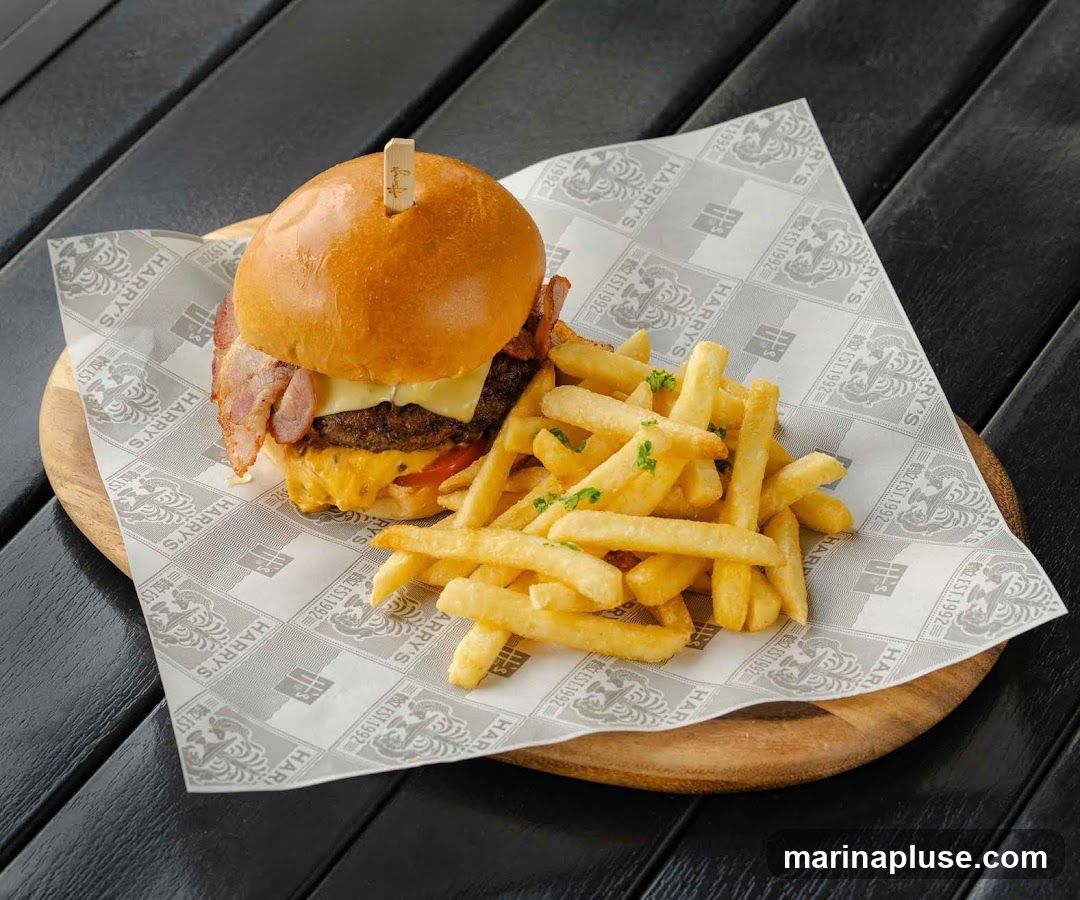A close-up shot of a burger and fries served on a wooden board with branded paper. The burger has a brioche bun, bacon, cheese, and tomato. The fries are golden brown and sprinkled with parsley. The paper has a repeating pattern of the text "HARRY'S EST. 1992" and a tiger logo.