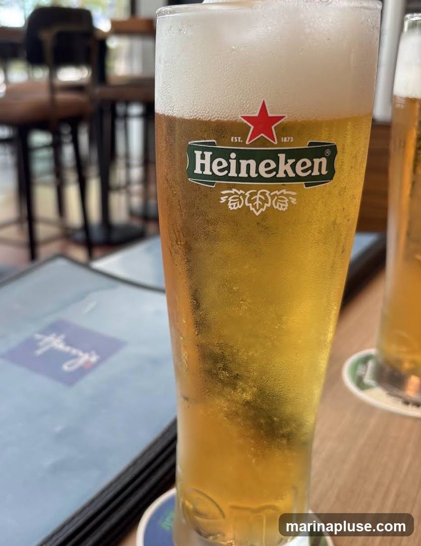 A close-up shot of a glass of Heineken beer with a foamy head. In the background, a menu with a blue cover and a logo is visible, along with blurred seating and tables, suggesting an indoor dining or bar setting. Another glass of beer is partially visible to the right.