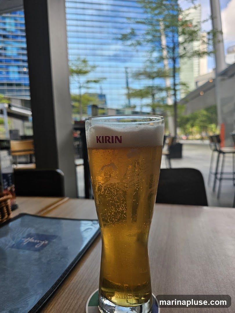 A tall glass of Kirin beer with a foamy head sits on a wooden table. To the left, a menu is visible, with the name 'Harry's' partially obscured. In the background, a modern building with a glass facade reflects the sky and trees.