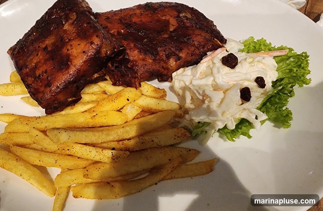 A plate of barbecued ribs served with french fries and coleslaw. The ribs are dark brown and appear to be glazed. The french fries are golden brown and seasoned. The coleslaw is creamy and topped with raisins. A lettuce leaf garnishes the coleslaw.