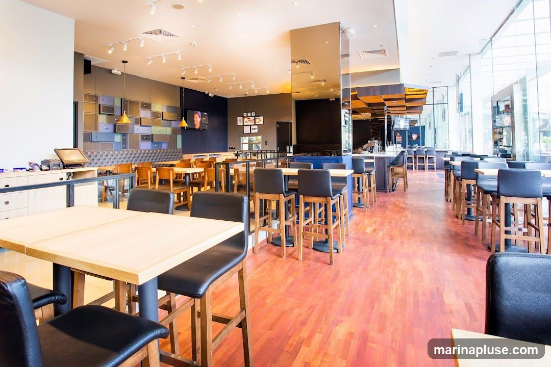 The image shows the interior of a modern restaurant with a bar area and dining tables. The decor features a wall with colorful drawers, a long bar with stools, and wooden tables with black chairs. Large windows allow natural light to fill the space.