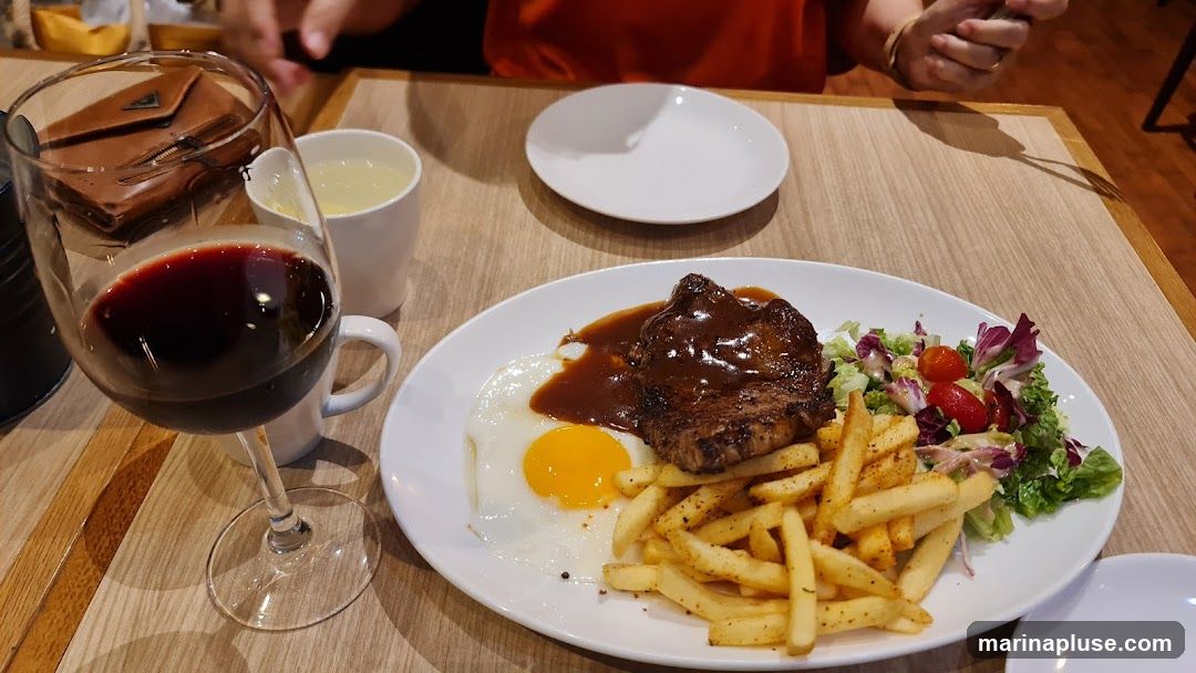 A plate of steak, a fried egg, french fries, and a side salad with cherry tomatoes and lettuce. A glass of red wine and a cup of a light-colored beverage are also on the table. A brown wallet is visible in the wine glass.