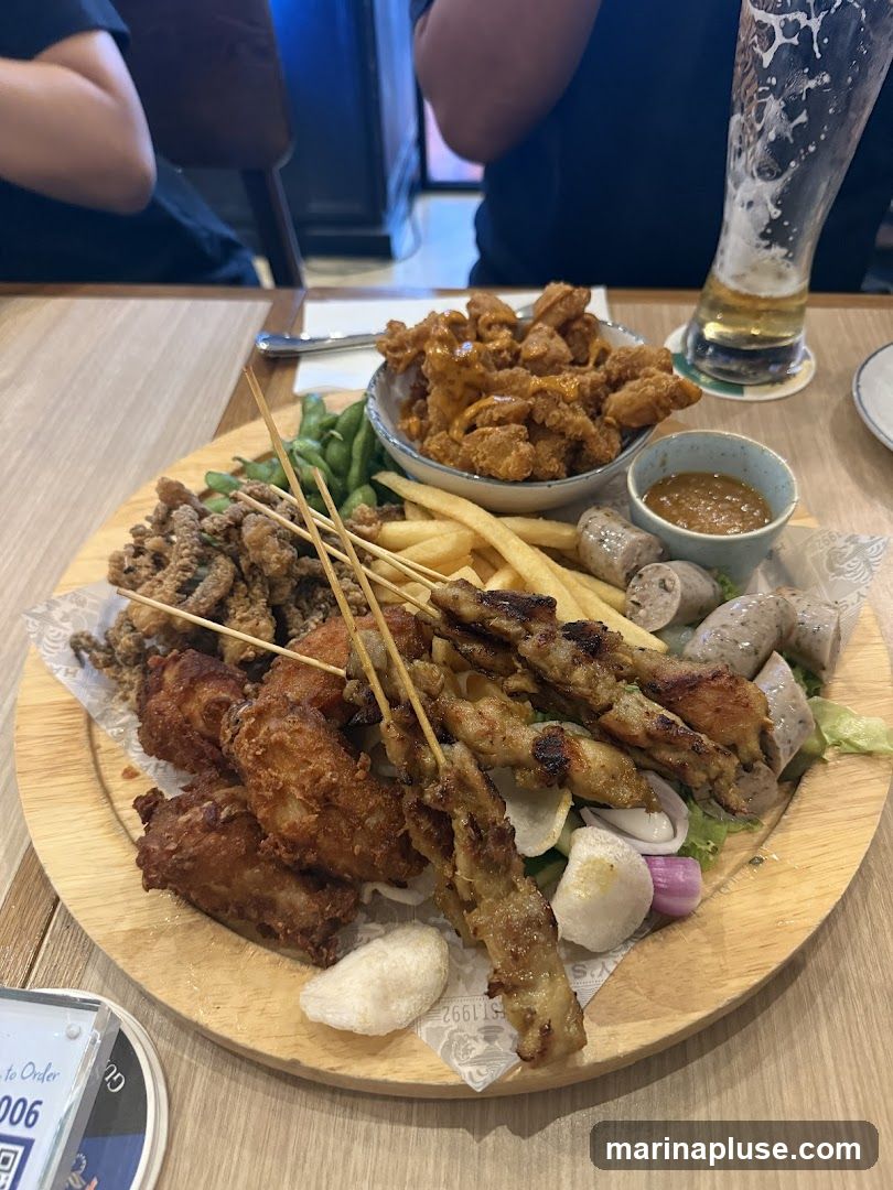 A wooden platter filled with various fried appetizers, including chicken wings, satay skewers, french fries, sausages, edamame, and a small bowl of dipping sauce. A glass of beer is visible in the background.