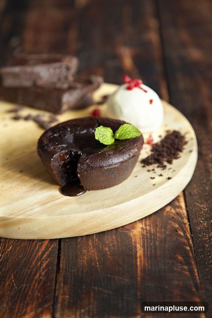 A close-up shot of a chocolate lava cake with a scoop of vanilla ice cream. The lava cake is oozing molten chocolate and is garnished with mint leaves. There are also some chocolate brownies and chocolate crumbs on the wooden serving board. The background is a dark wooden surface.
