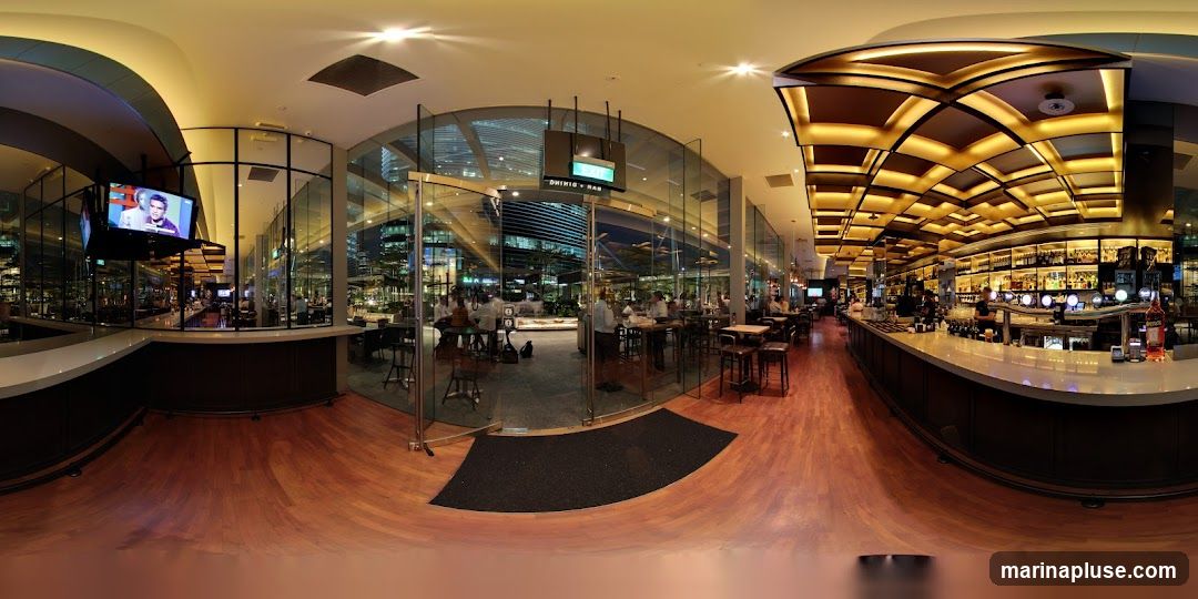A panoramic view of a modern bar and dining area with a long, illuminated bar stocked with bottles. Seating areas with tables and chairs are visible, as well as a television screen displaying a program. The interior features warm lighting and wooden floors, with large glass windows offering a view of the city at night.