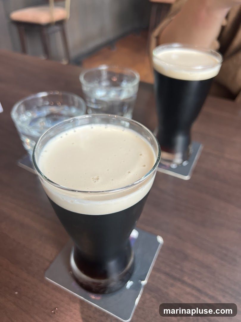 Two glasses of dark beer with foamy heads and two glasses of water are on a wooden table. The glasses of beer are on coasters. In the background, a chair and a brown paper bag are visible.