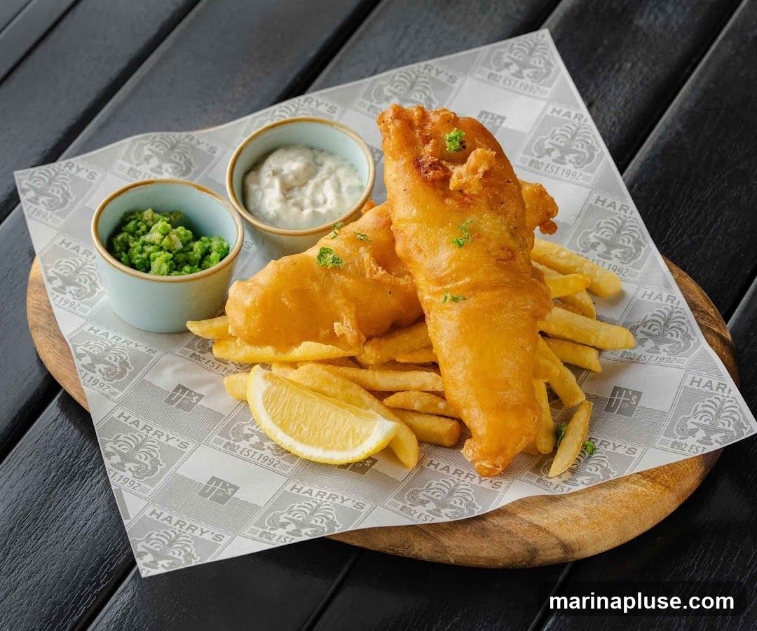 A plate of fish and chips served with mushy peas and tartar sauce, garnished with lemon and parsley. The food is presented on a wooden board lined with paper featuring the Harry's logo and "EST. 1992".