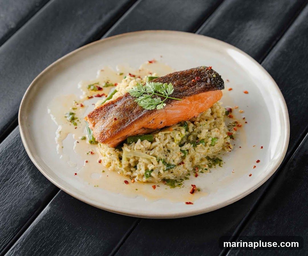 A close-up shot of a plate of food featuring a piece of salmon fillet served over a bed of rice with green vegetables. The dish is garnished with fresh herbs and sprinkled with red pepper flakes. The plate is set on a dark wooden surface.