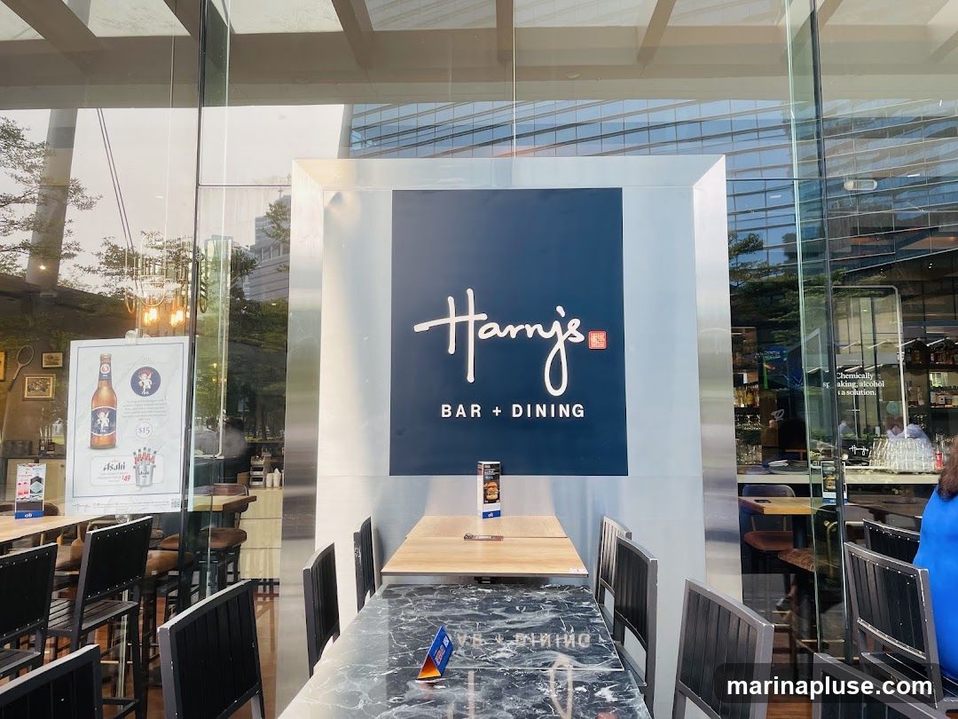 The exterior of Harry's Bar + Dining shows a large blue sign with white lettering that reads "Harry's BAR + DINING". The entrance is framed by a metallic border. Through the glass windows, tables and chairs are visible, along with a bar area. A poster advertising Asahi beer is displayed on the left side of the entrance.