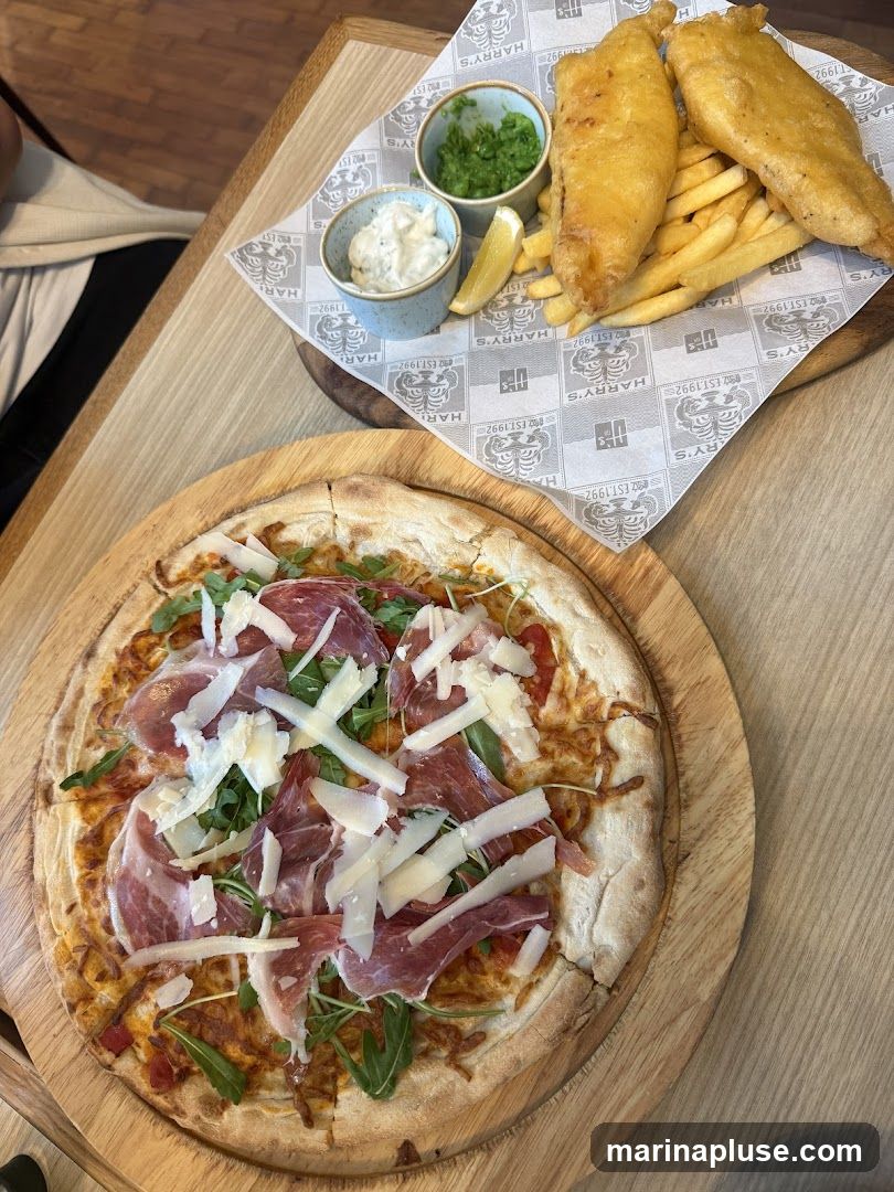A close-up overhead shot of a pizza and a serving of fish and chips on a wooden table. The pizza is topped with prosciutto, arugula, and shaved parmesan cheese. The fish and chips are served with mushy peas, tartar sauce, and a lemon wedge on branded paper. The branding on the paper indicates it is from "Harry's Est. 1992".