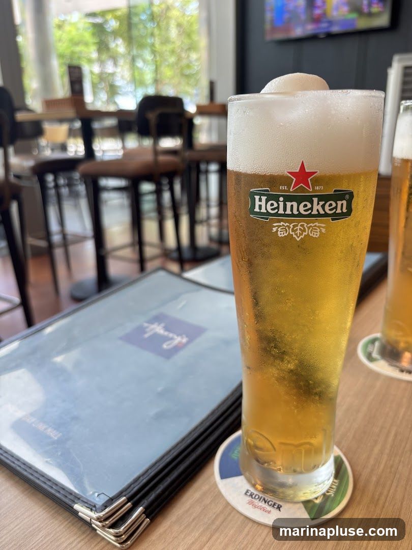 A close-up of a glass of Heineken beer with a foamy head sits on a table next to a menu. In the background, blurred tables and chairs suggest a restaurant or bar setting. Another glass of beer and a coaster are also visible.