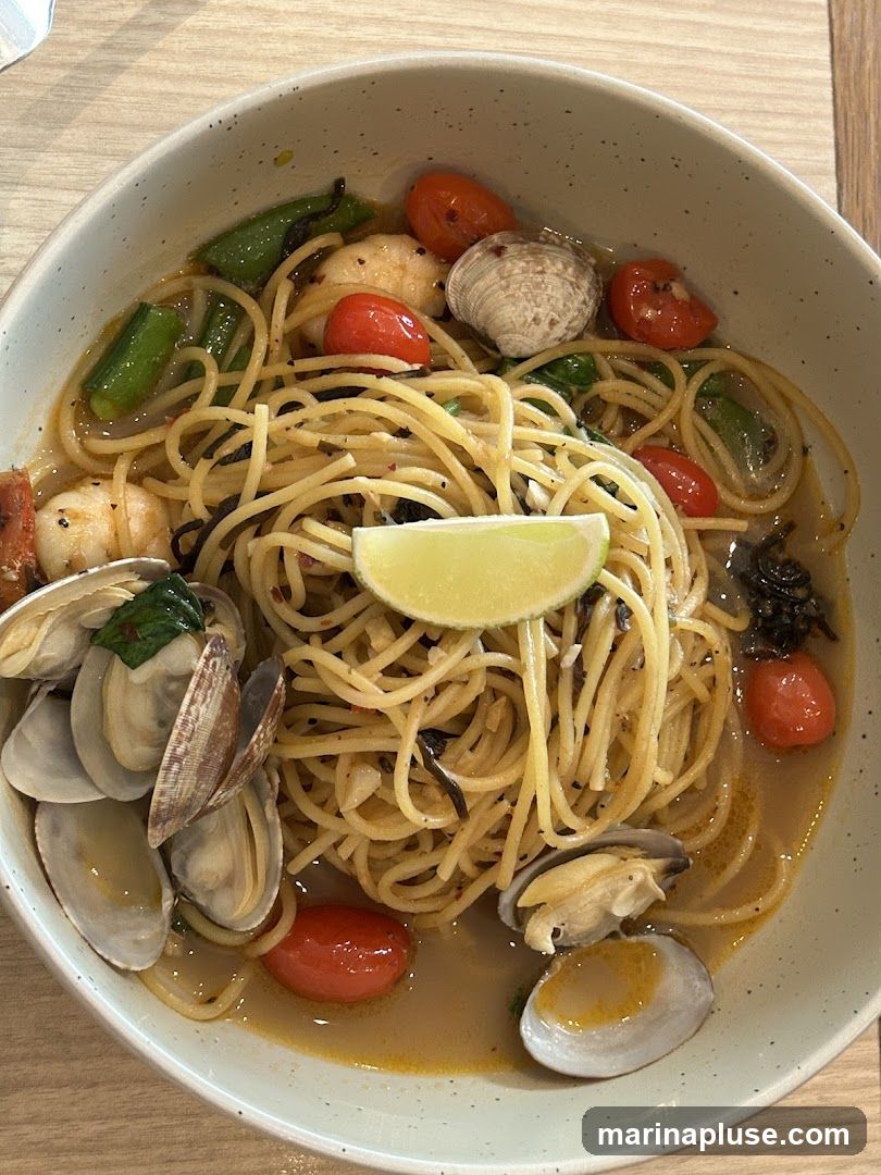A close-up shot of a bowl of spaghetti with clams, shrimp, cherry tomatoes, and green beans. The pasta is coated in a light, savory sauce and garnished with a slice of lime.