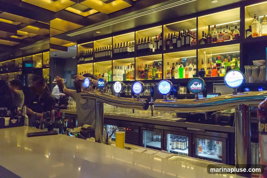 A well-lit bar interior featuring a polished counter in the foreground. Behind the counter, shelves are stocked with a variety of bottles and spirits, illuminated by warm lighting. Several beer taps with illuminated logos are prominent, including Asahi, Carlsberg, and Thirstyale. A glass of yellow liquid, likely beer, sits on the counter. In the background, patrons and staff are visible, suggesting a lively atmosphere. The ceiling has a distinctive geometric design with gold accents.