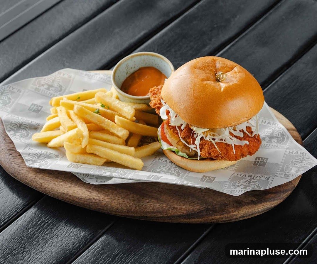 A close-up shot of a crispy chicken burger served with a side of golden french fries and a small bowl of dipping sauce. The burger is on a bun with shredded lettuce, tomato, and a thick chicken patty. The food is presented on a wooden board lined with paper featuring the "Harry's Est. 1992" logo.