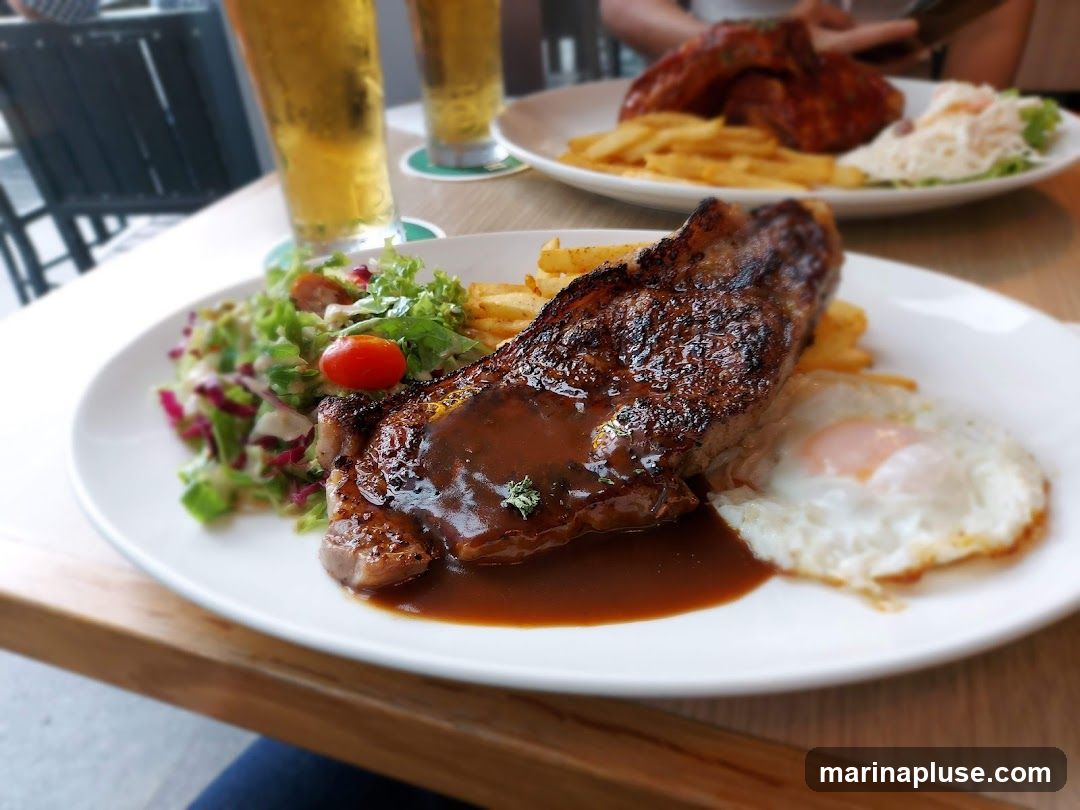 A close-up shot of a steak dinner served on a white plate. The steak is topped with a rich brown sauce and accompanied by french fries, a side salad with a cherry tomato, and a fried egg. In the background, two glasses of beer and another plate with ribs and fries are visible.
