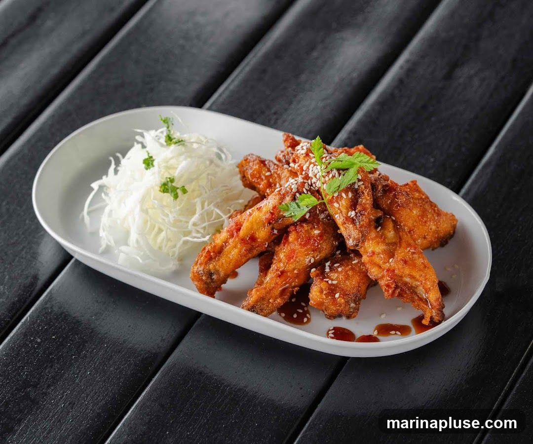 A white oval plate holds several fried chicken wings coated in a glossy sauce and sprinkled with sesame seeds and parsley. A pile of shredded white cabbage is served on the side. The plate rests on a dark, textured surface made of wooden planks.
