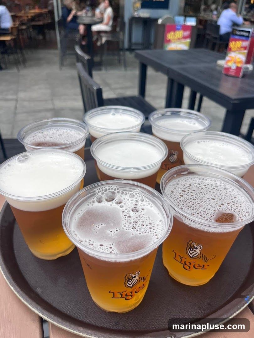 A tray holding eight plastic cups of Tiger beer, each with a foamy head. The cups have the Tiger logo visible. In the background, an outdoor seating area of a restaurant or bar is visible with patrons seated at tables. There are also some promotional signs on tables in the background.