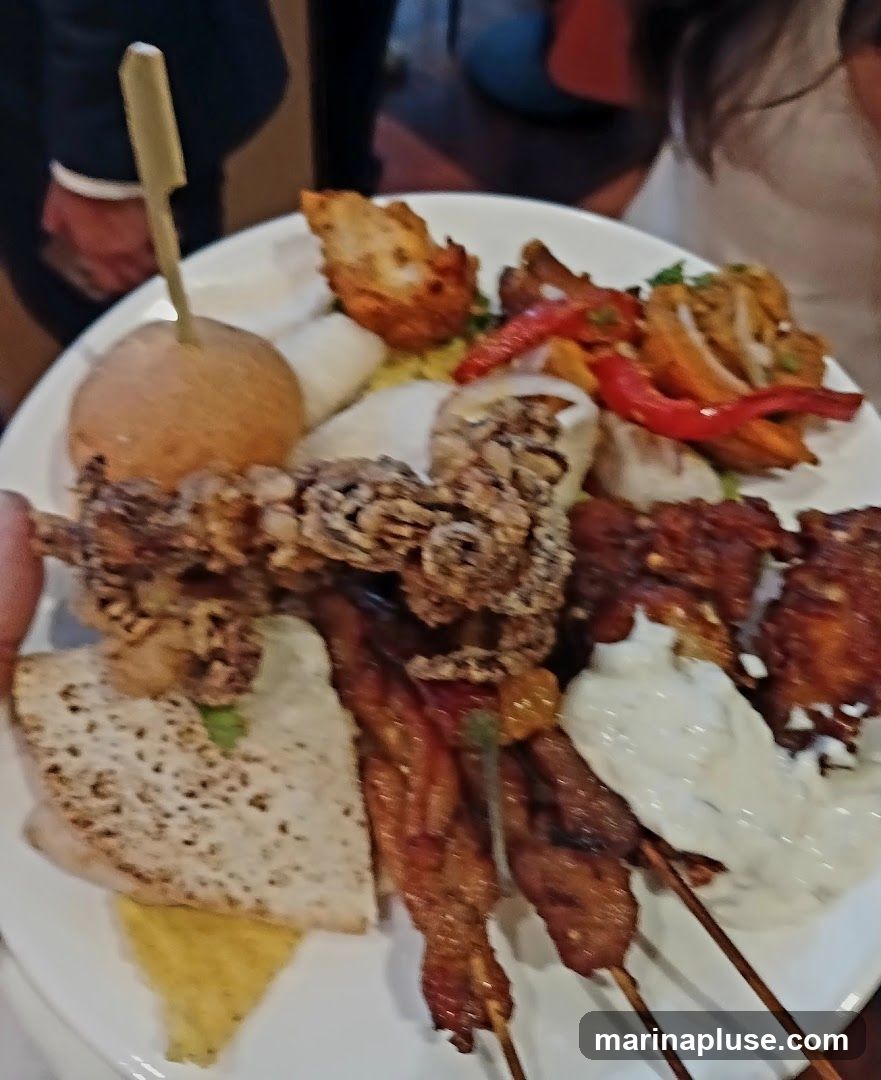 A white plate filled with various food items, including what appears to be fried chicken wings, skewers of meat, a small bun with a skewer, bread, and a dollop of white sauce. There are also some red peppers and other unidentifiable fried items. The plate is held by a person, and other people are visible in the background.