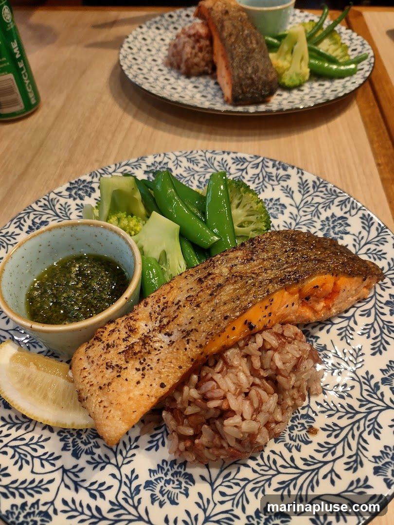 A close-up shot of a plate of food featuring a piece of pan-seared salmon served with brown rice and a side of steamed broccoli and snap peas. A small bowl of green sauce and a lemon wedge are also on the plate. Another plate with a similar meal is visible in the background. A can with the text "RECYCLE ME" is on the left side of the image.