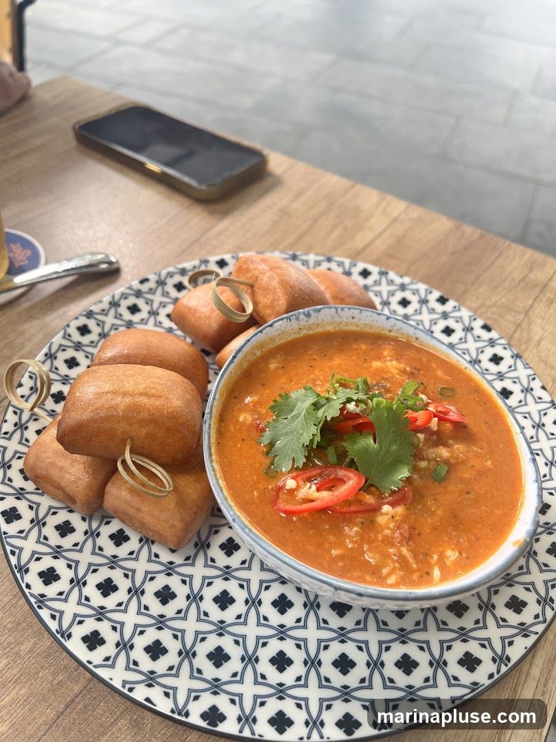 A close-up shot of a bowl of orange soup, garnished with cilantro and sliced red chilies, served with fried bread on a decorative plate. A smartphone is visible in the background on a wooden table.