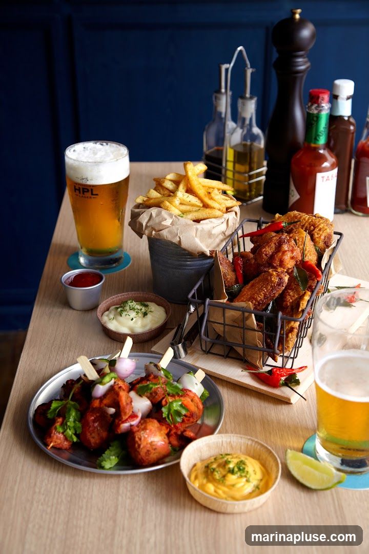 A table laden with various appetizers and drinks, including chicken skewers, fried chicken wings, french fries, dips, and two glasses of beer. Condiments and a pepper mill are also visible in the background.