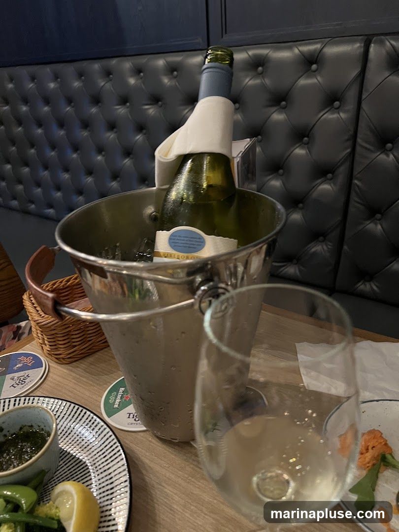 A bottle of white wine in an ice bucket sits on a wooden table, next to a glass of white wine. A plate with green vegetables and a lemon wedge is also visible, along with coasters and a woven basket. The background features a dark, tufted upholstered bench.
