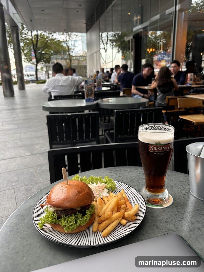 A close-up view of a burger and fries served on a plate, with a glass of dark beer next to it. The meal is placed on an outdoor table, with blurred background showing people dining at other tables and the exterior of a building. The beer glass has a Kilkenny Draught logo.