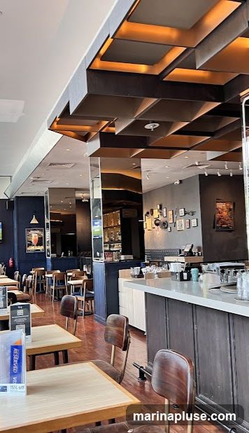 The image shows the interior of a restaurant with wooden tables and chairs. A bar area is visible on the right side of the image, with shelves stocked with bottles behind it. The ceiling features a modern design with recessed lighting. Framed pictures and artwork adorn the walls. A sign on a table in the foreground indicates "RESERVED" and "15% off".