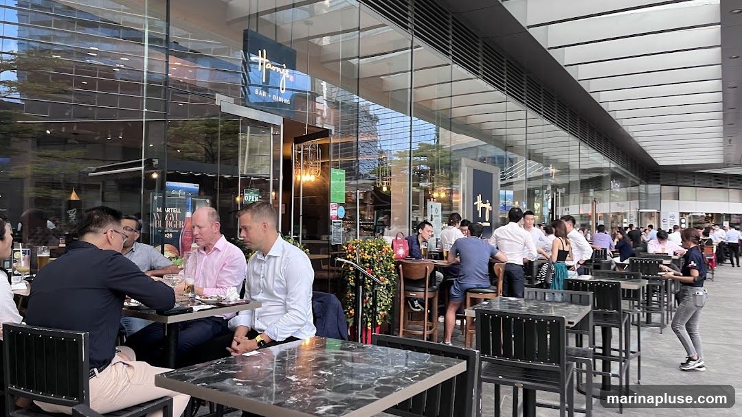 A street-level view of Harry's Bar + Dining, a restaurant with a modern glass facade. People are seated at tables both inside and on the outdoor patio. The restaurant's sign is visible above the entrance.