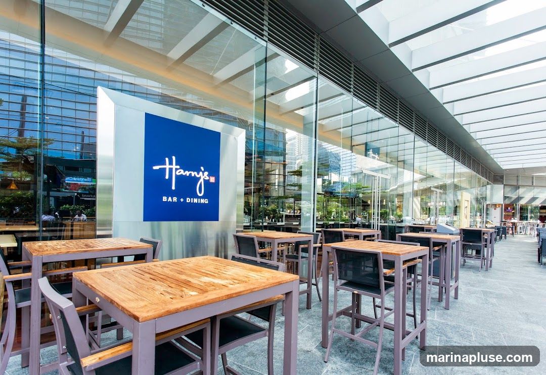 The exterior of a modern bar and dining establishment with a prominent blue sign featuring the 'Harry's' logo and text 'BAR + DINING'. The storefront has large glass windows and an outdoor seating area with wooden tables and chairs.