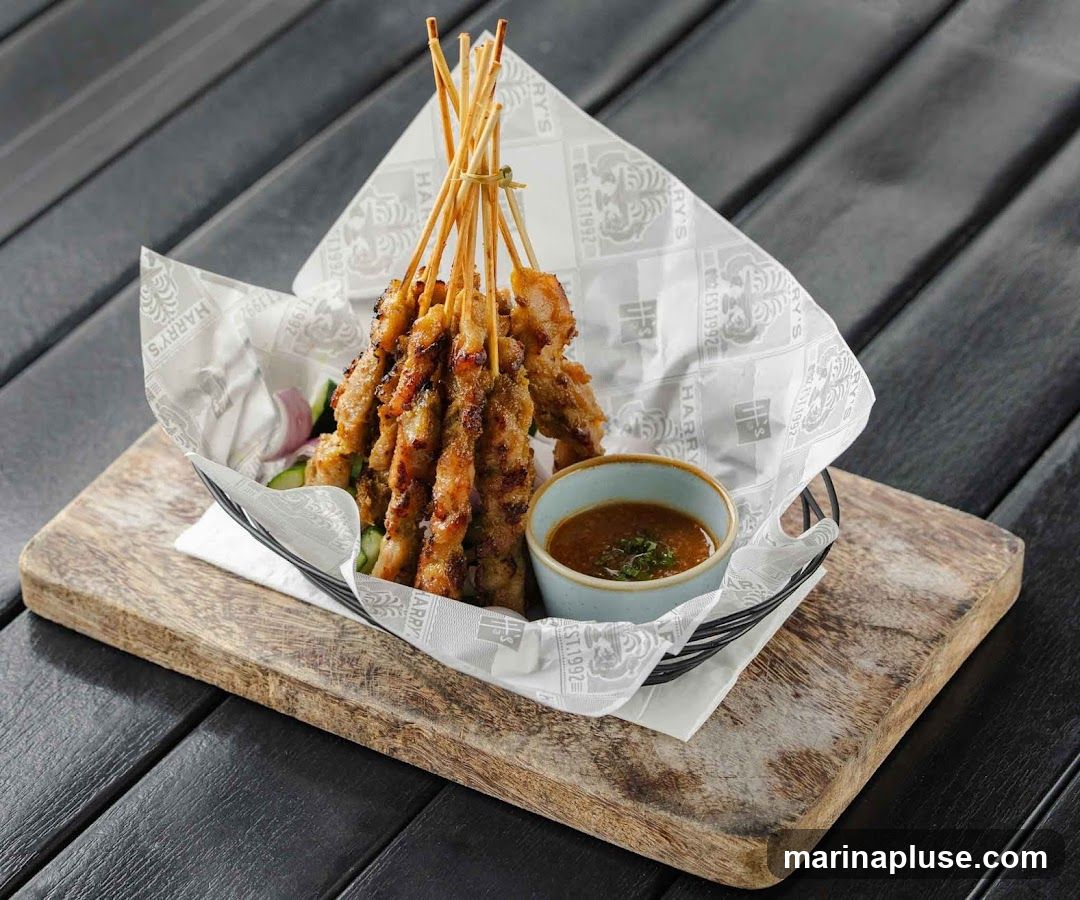 A basket of grilled chicken satay skewers served with a side of peanut sauce, cucumber, and red onion slices. The skewers are presented on a wooden board with branded paper lining the basket. The paper has the "HARRY'S EST. 1992" logo repeated.
