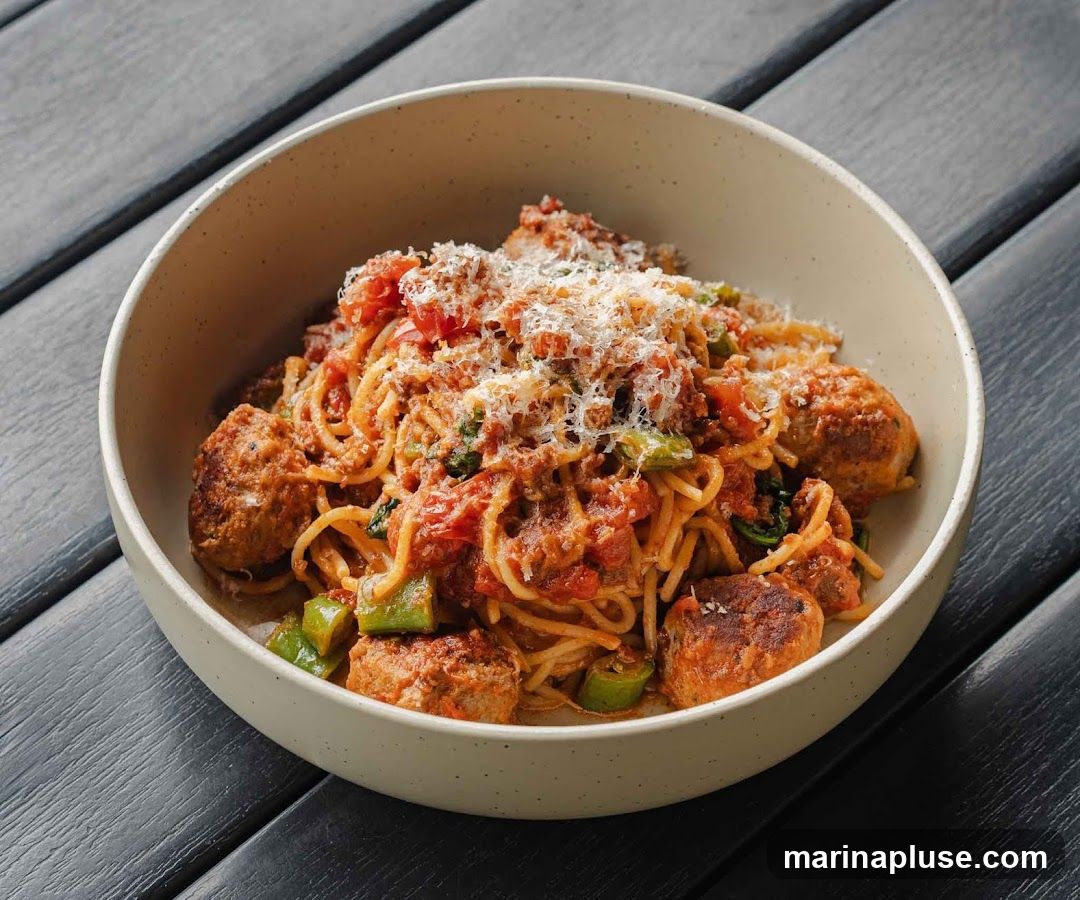 A close-up shot of a bowl of spaghetti and meatballs. The pasta is coated in a rich tomato sauce with visible chunks of tomatoes and green peppers. Several large, browned meatballs are nestled amongst the spaghetti. The dish is generously topped with grated Parmesan cheese. The bowl is light-colored and sits on a dark, slatted surface.