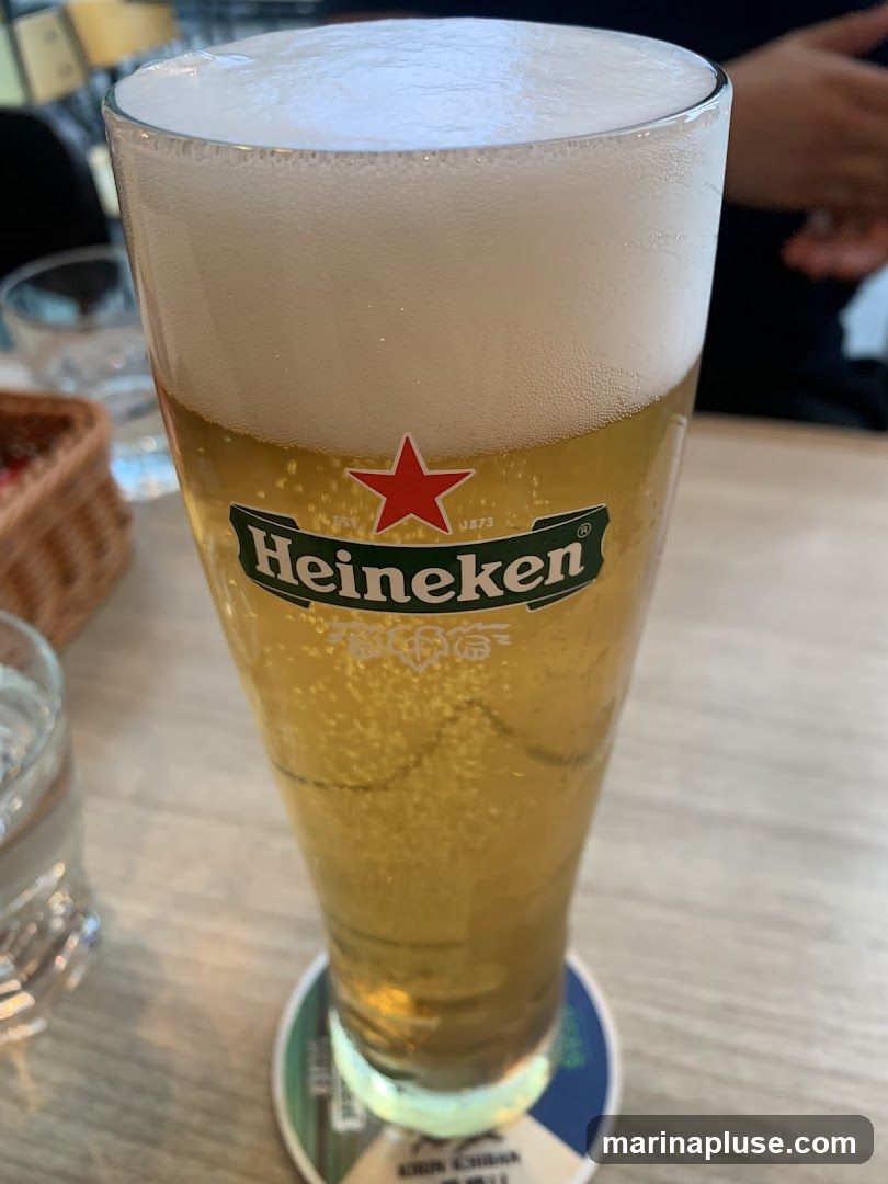 A tall glass of Heineken beer with a thick foamy head sits on a table. The Heineken logo with a red star is visible on the glass. Bubbles are visible in the beer. In the background, a wicker basket and a glass of water can be seen.