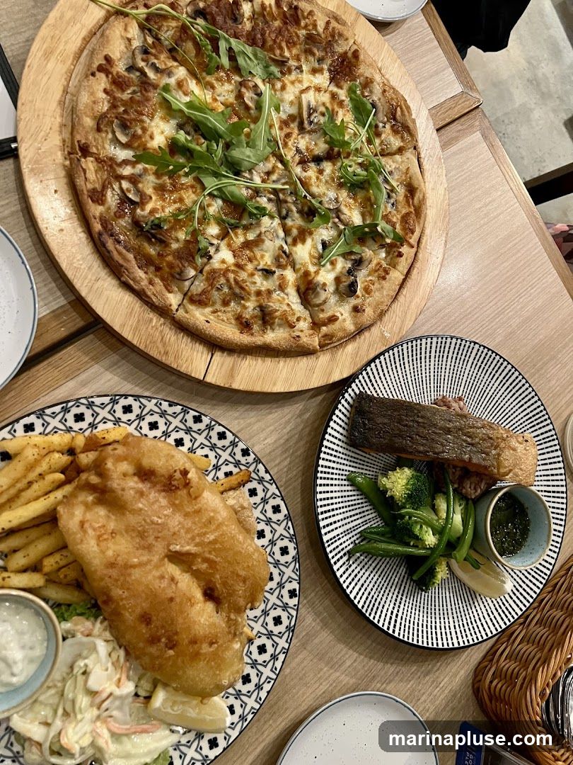 A top-down view of a table laden with food. In the center is a large pizza topped with mushrooms and arugula. To the left, a plate of fish and chips with coleslaw and a side of sauce. To the right, a piece of salmon served with rice and steamed vegetables, accompanied by a small bowl of green sauce.