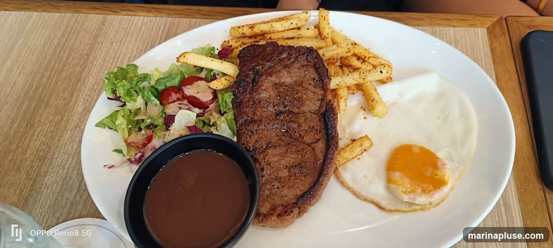 A plate of food featuring a steak, french fries, a side salad with cherry tomatoes and dressing, a fried egg, and a small bowl of sauce. The meal is served on a white plate on a wooden table.