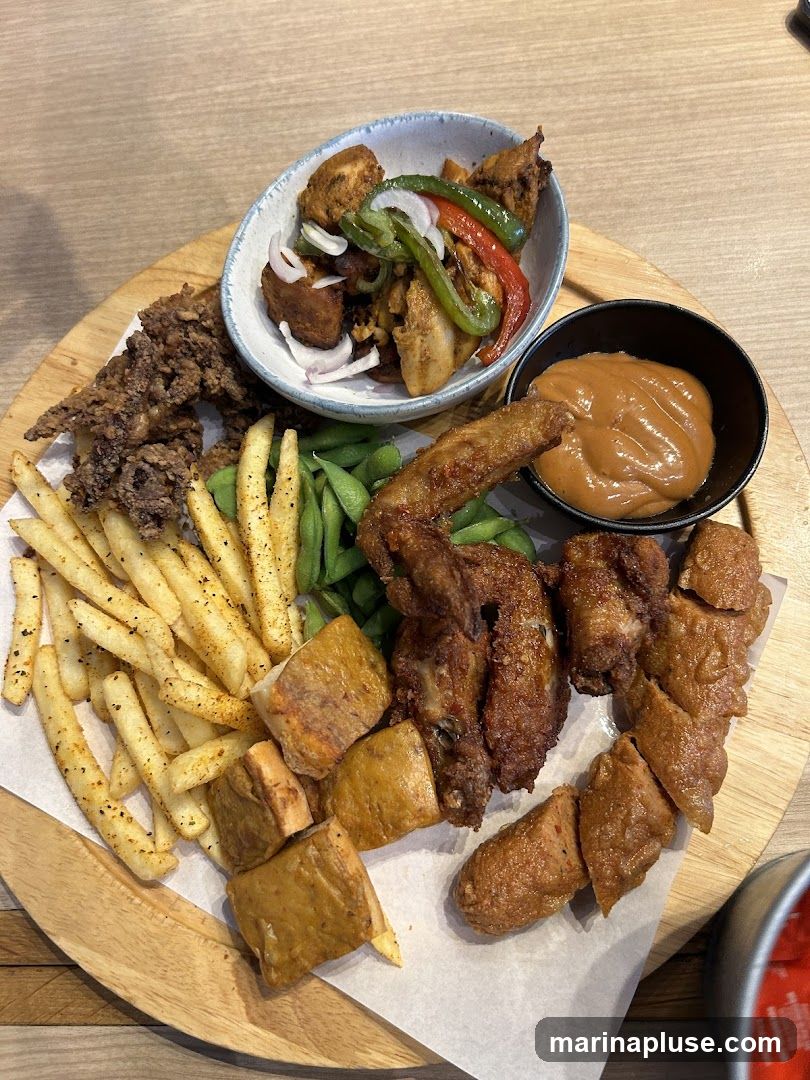A wooden platter filled with various fried items, including french fries, chicken wings, fried calamari, and other assorted fried pieces. There is also a small bowl of edamame and a bowl of dipping sauce. A separate bowl contains fried chicken pieces with bell peppers and onions.
